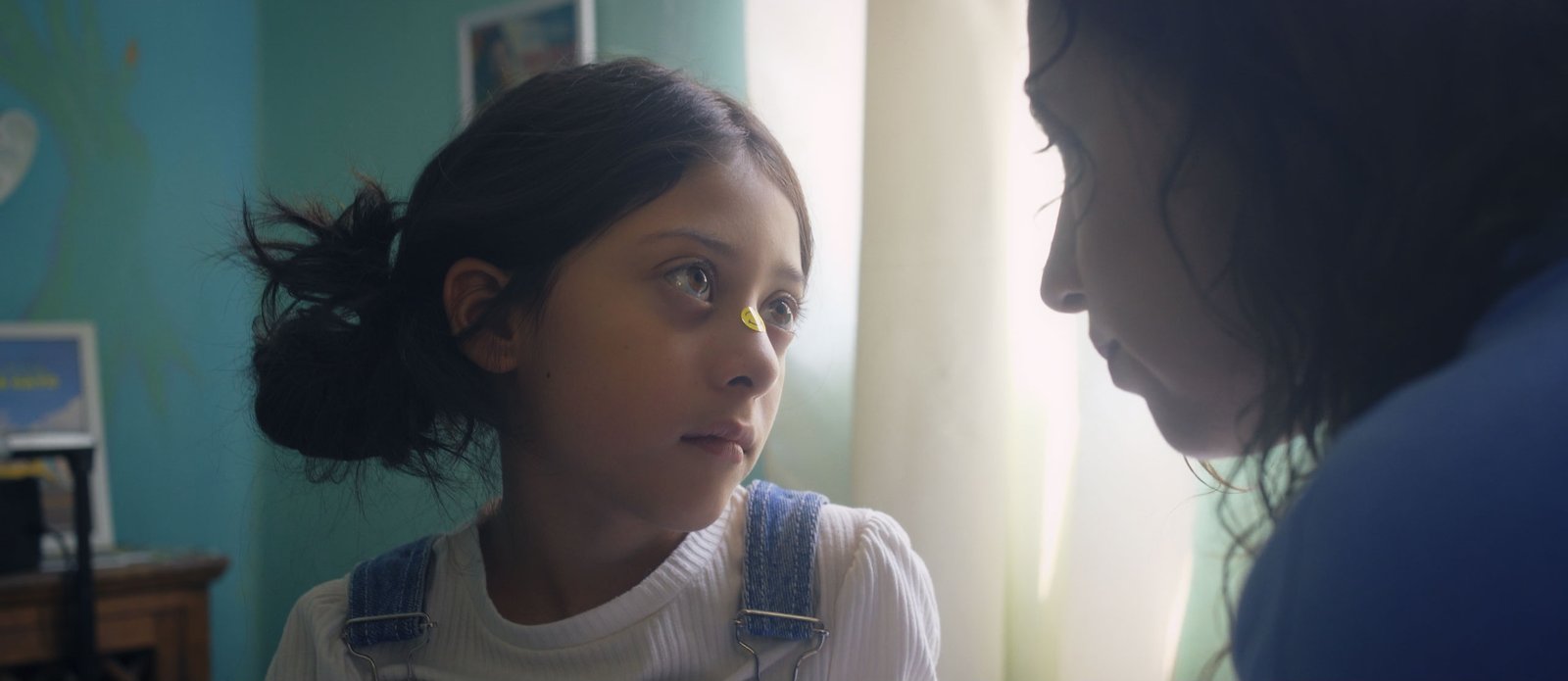 Fotograma de Para que no se te olvide: una niña con una estampa en la nariz mira a su madre junto a una ventana